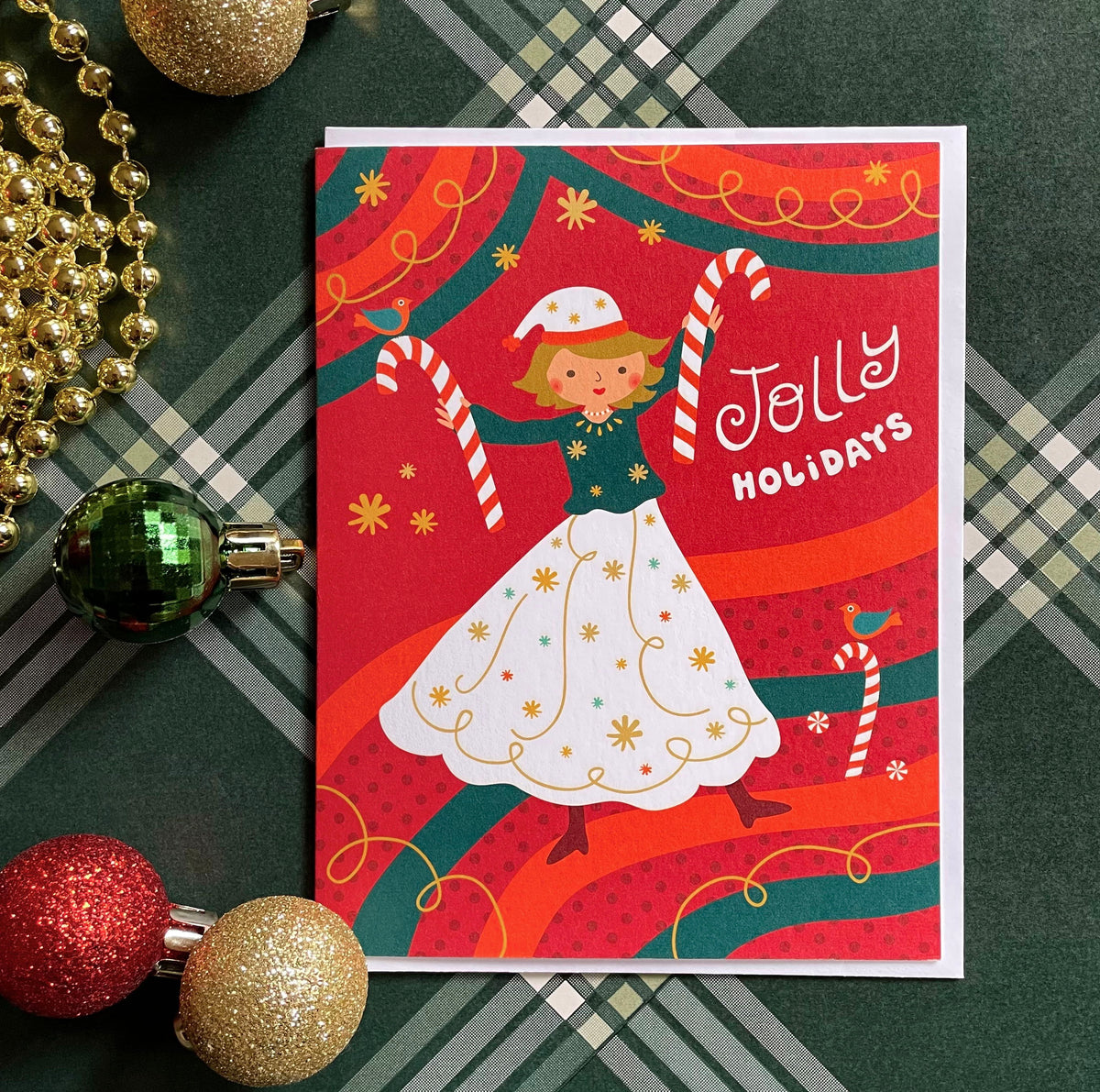 Jolly Gal folded paper holiday card featuring a dancing woman in a white dress and Santa hat holding candy canes against a bold red and green swirling background with colorful birds and the greeting "Jolly Holidays." Shown with a white envelope and a green plaid table covering surrounded by glittery Christmas tree ornaments.