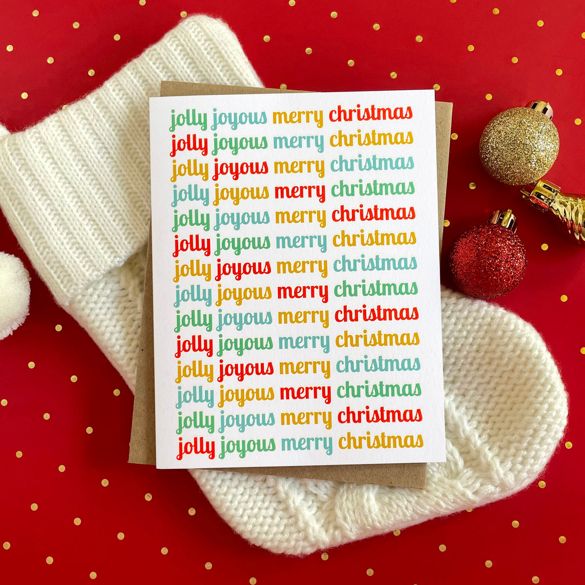 Jolly Joyous folded paper holiday card featuring the words "jolly joyous merry christmas" repeated in rotating red, teal, gold and green script across a white background. Card is paired with a kraft envelope and sits atop a cream stocking on a red polka dot table covering.