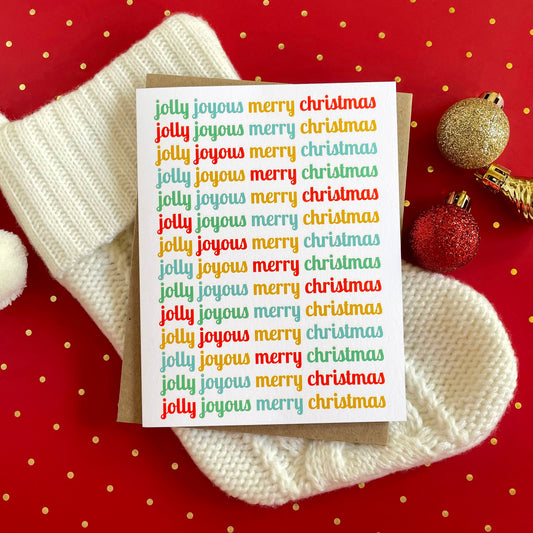 Jolly Joyous folded paper holiday card featuring the words "jolly joyous merry christmas" repeated in rotating red, teal, gold and green script across a white background. Card is paired with a kraft envelope and sits atop a cream stocking on a red polka dot table covering.