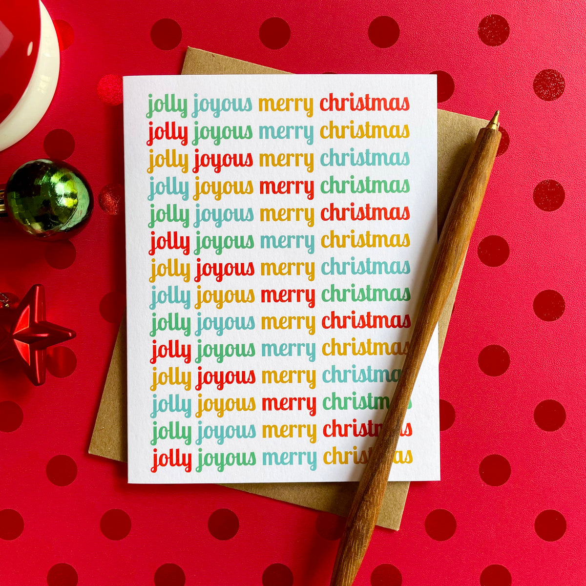 Jolly Joyous folded paper holiday card featuring the words "jolly joyous merry christmas" repeated in rotating red, teal, gold and green script across a white background. Card is paired with a kraft envelope and sits atop a red polka dot table covering.