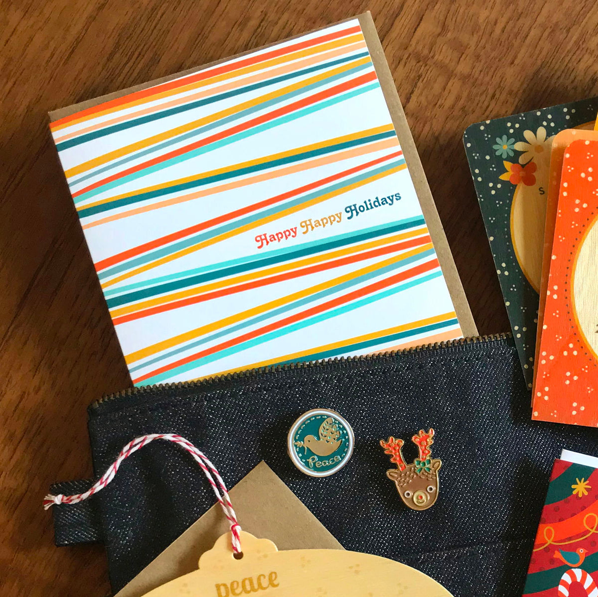 Happy Stripes folded paper holiday card featuring sweeping diagonal stripes in orange, teal, gold, peach and slate blue on a white background with the greeting "Happy Happy Holidays." Shown with a kraft envelope. Also pictured are a reindeer and peace dove enamel pin.