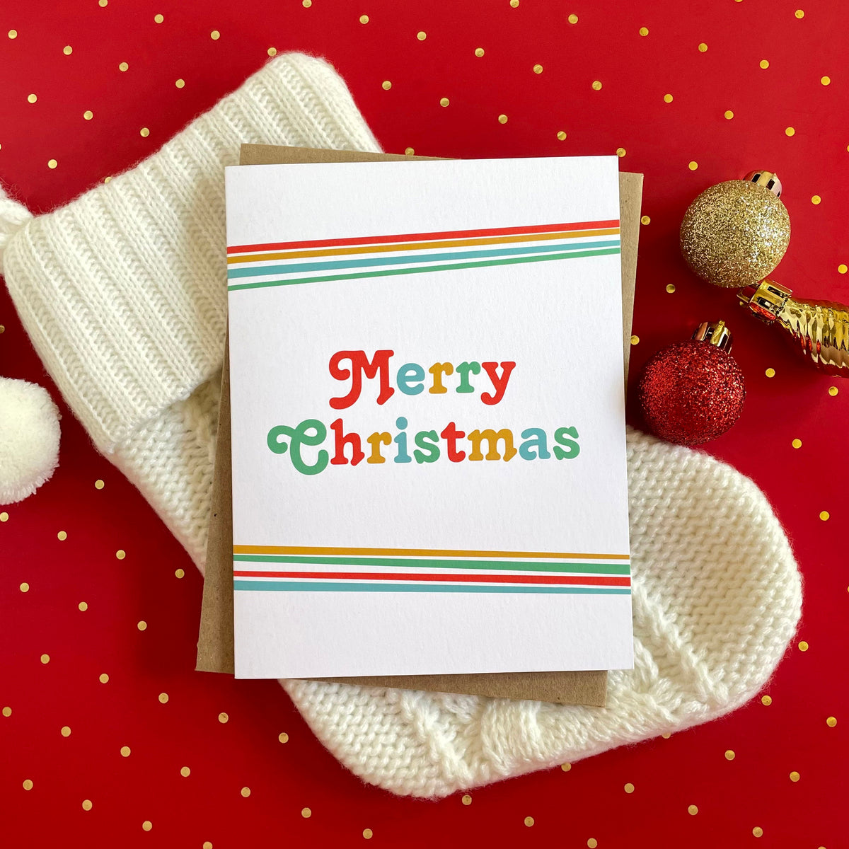 Big Christmas folded paper holiday card featuring bold multicolored "Merry Christmas" lettering in red, green, teal and gold on a white background with rainbow stripes along the top and bottom edges. Card is paired with a kraft envelope and sits atop a cream stocking on a red polka dotted table covering.