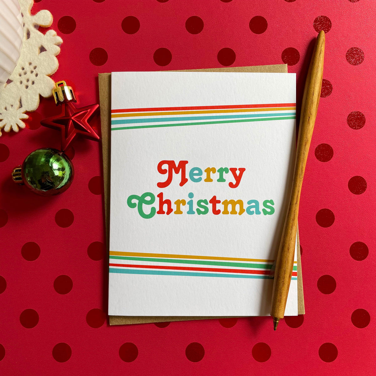 Big Christmas folded paper holiday card featuring bold multicolored "Merry Christmas" lettering in red, green, teal and gold on a white background with rainbow stripes along the top and bottom edges. Card is paired with a kraft envelope and sits atop a red polka dotted table covering.