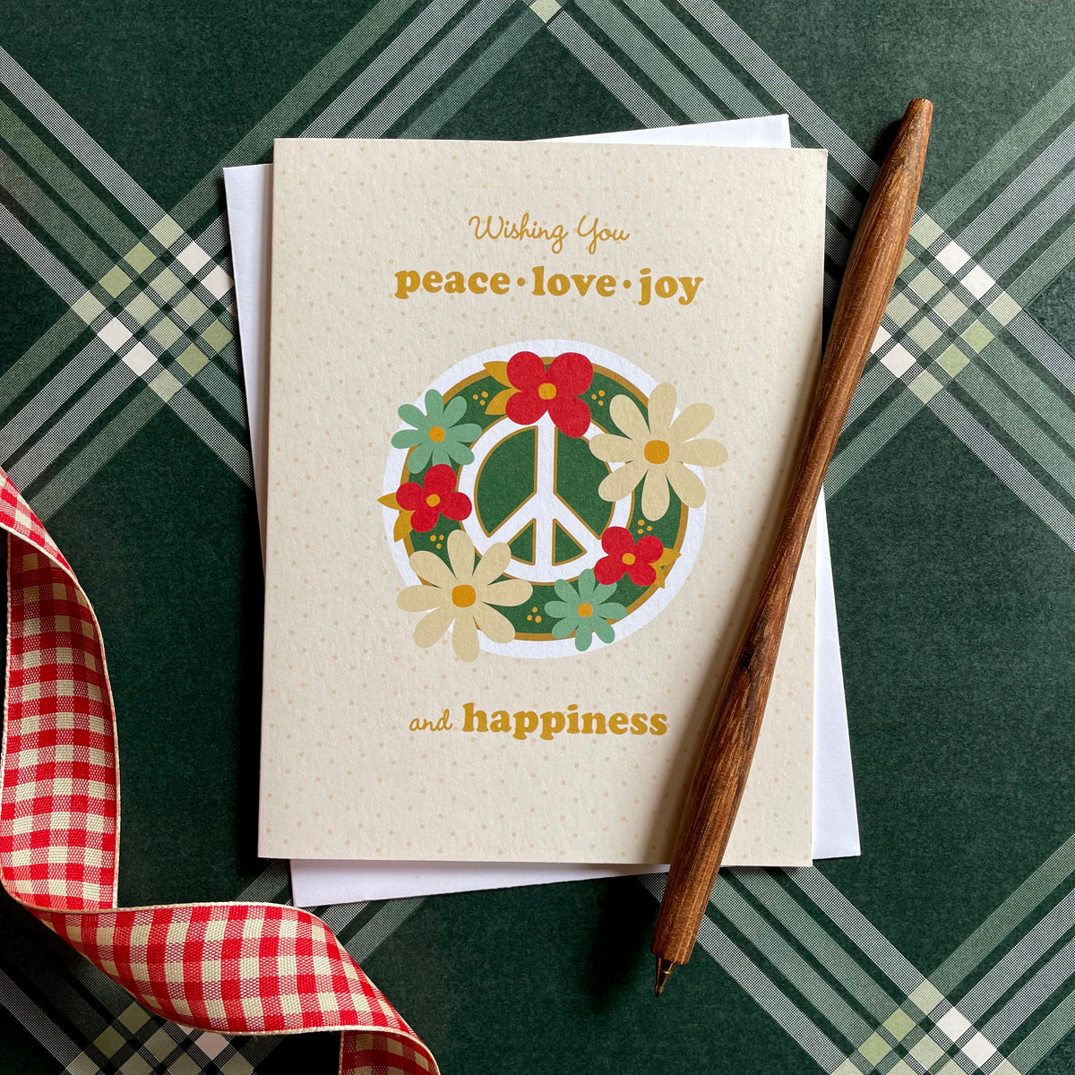 Blooming Peace Cream folded paper holiday card featuring a green peace sign surrounded by red, cream and teal flowers on a soft cream dotted background with the greeting "Wishing You peace, love, joy and happiness." Card is paired with a white envelope and sits atop a green plaid surface.