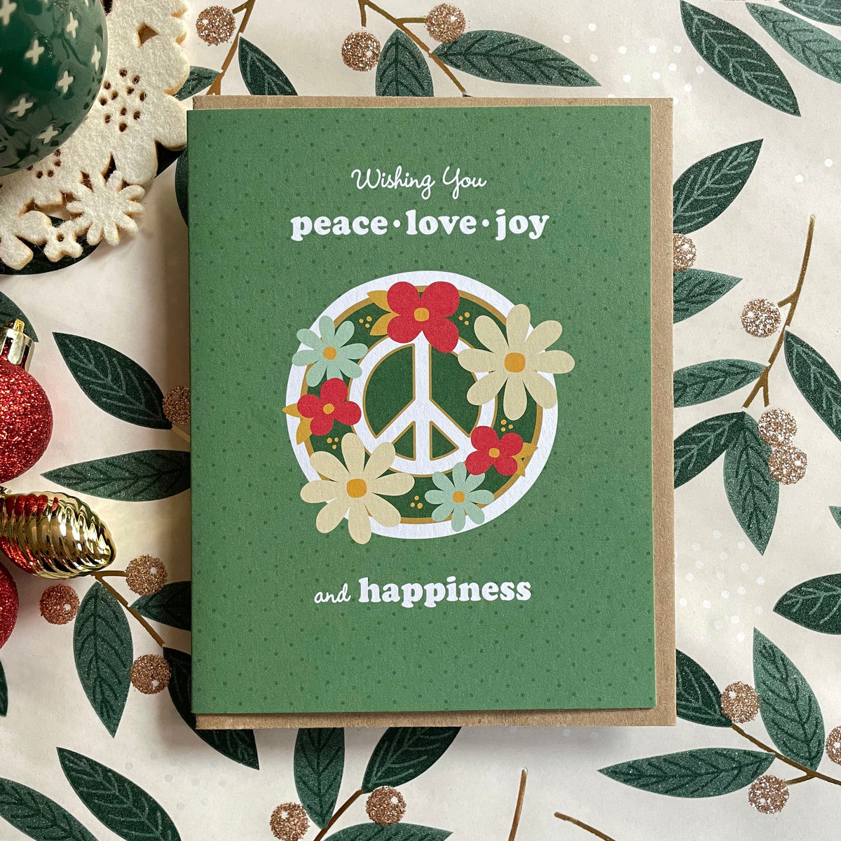Blooming Peace Green folded paper holiday card featuring a green peace sign surrounded by red, cream and teal flowers on a soft green dotted background with the greeting "Wishing You peace, love, joy and happiness." Card is paired with a kraft envelope and sits atop a festive table decoration.