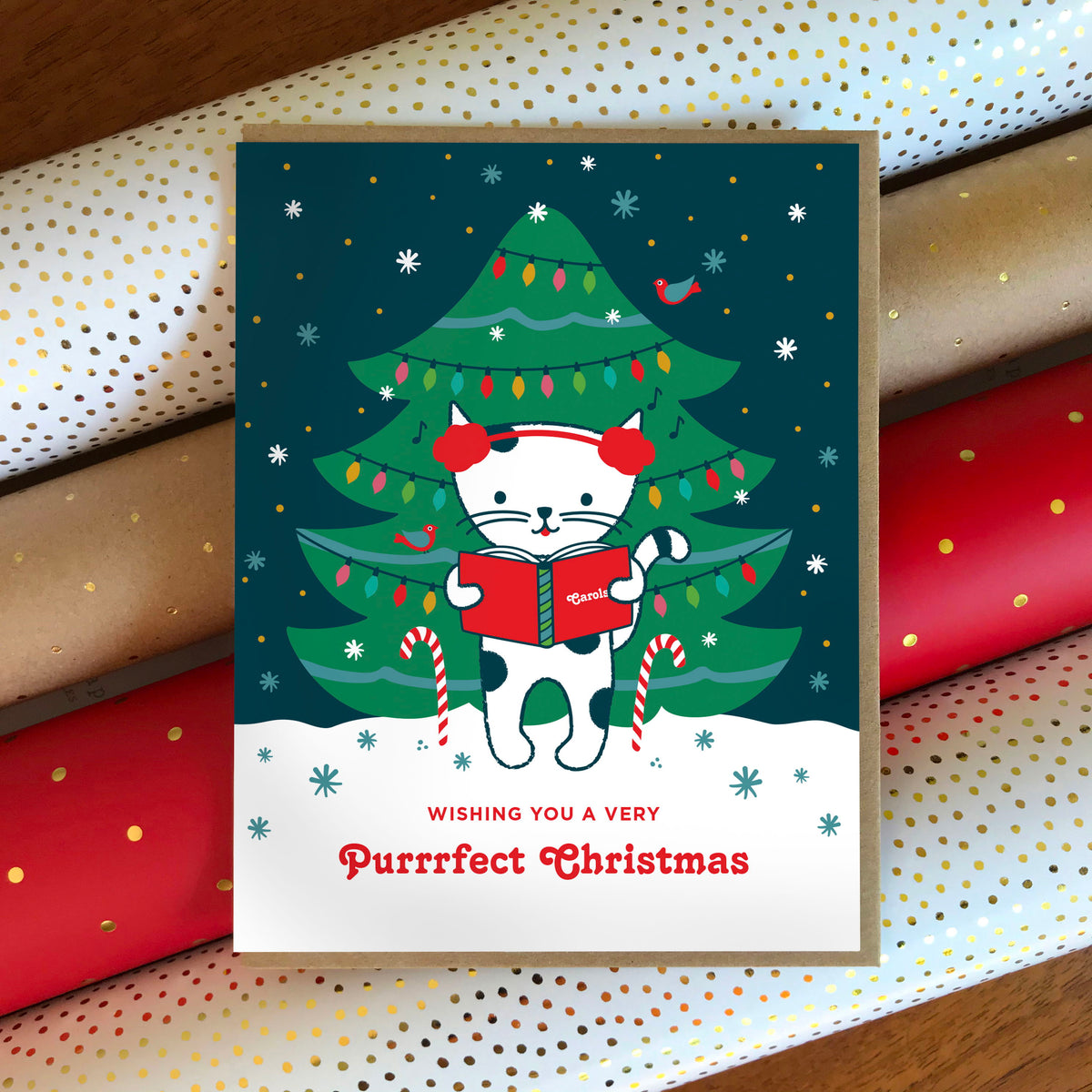 Caroling Kitty folded paper holiday card featuring a white cat in red earmuffs holding a carol book in front of a decorated Christmas tree with colorful lights, with the greeting "Wishing you a Very Purrrfect Christmas." Card is paired with a kraft envelope and sits atop rolls of festive wrapping paper.