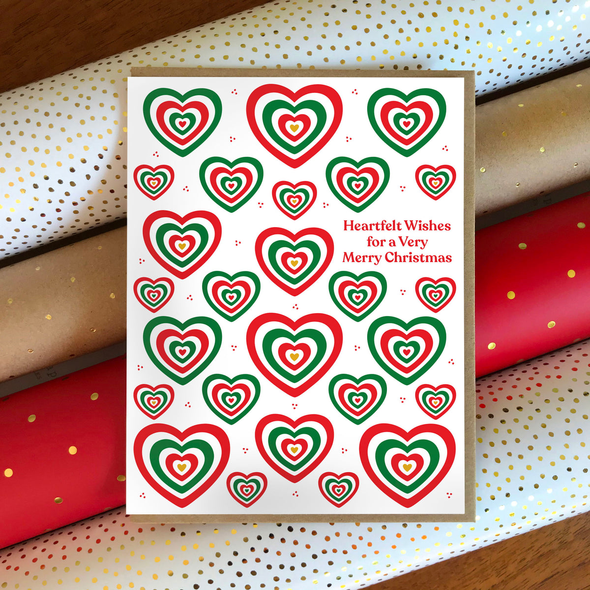 Heartfelt Wishes folded paper holiday card featuring bold red and green concentric hearts in various sizes scattered across a white background with the greeting "Heartfelt Wishes for a Very Merry Christmas." Card sits atop rolls of festive wrapping paper and is paired with a kraft envelope.