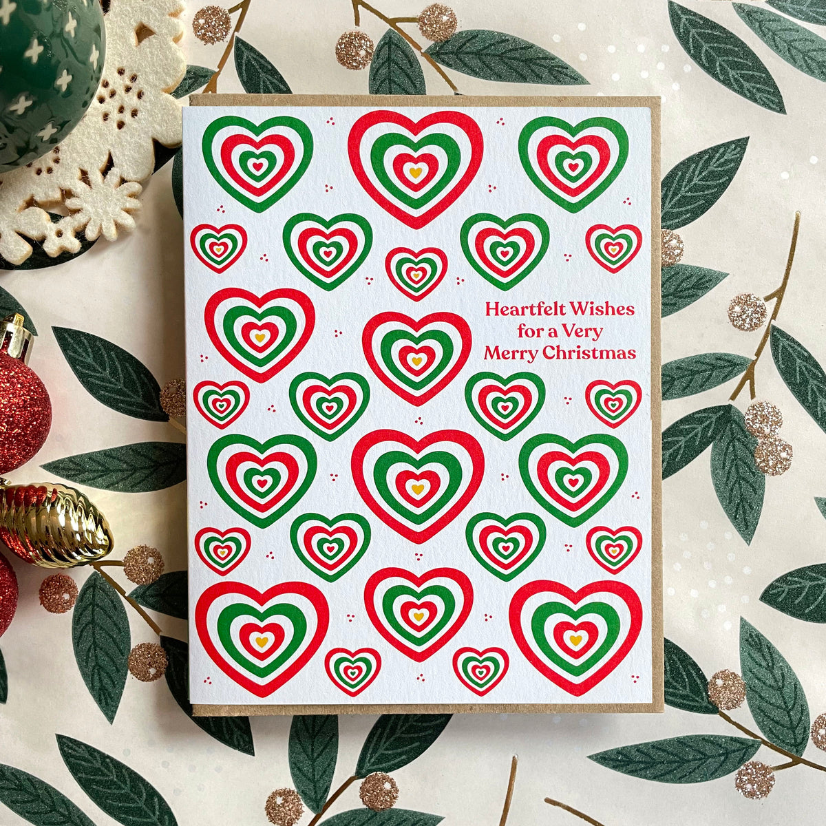 Heartfelt Wishes folded paper holiday card featuring bold red and green concentric hearts in various sizes scattered across a white background with the greeting "Heartfelt Wishes for a Very Merry Christmas." Card sits on a festive holly tablecloth and is paired with a kraft envelope.