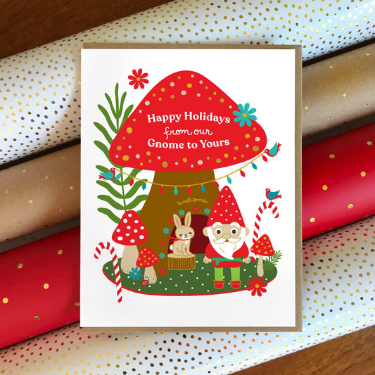 Gnome Home folded paper holiday card featuring a gnome and a bunny beside a large red mushroom house decorated with string lights, candy canes and colorful flowers, with the greeting "Happy Holidays from our Gnome to Yours." Card sits atop rolls of festive wrapping paper and is paired with a kraft envelope.