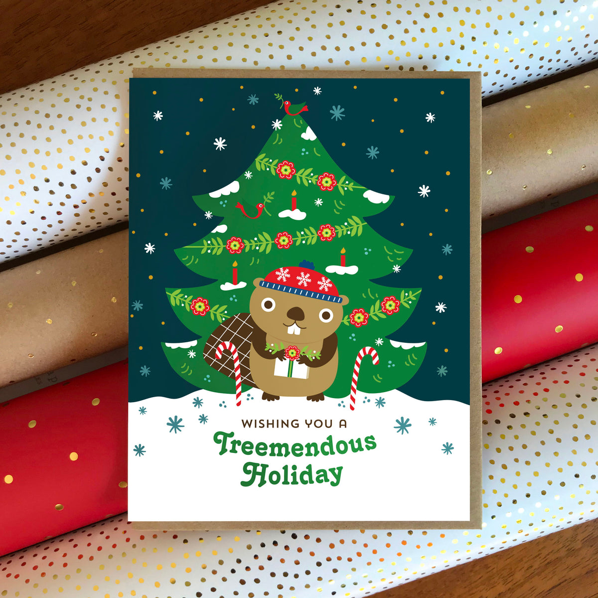Treemendous Holiday folded paper holiday card featuring a beaver in a red winter hat holding a gift in front of a decorated Christmas tree with red flowers, candles and a red bird on top, with the greeting "Wishing you a Treemendous Holiday." Card sits atop rolls of festive wrapping paper and is shown with a kraft envelope. 