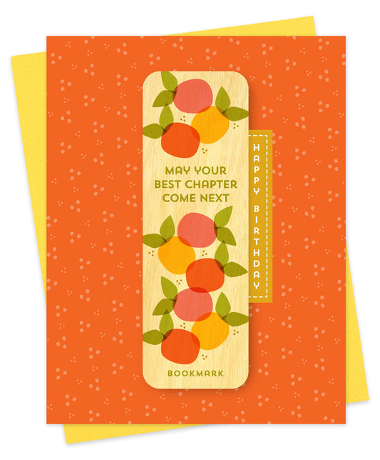 Folded birthday greeting card with a vibrant orange speckled background, Happy Birthday text and a removable birch wood bookmark featuring colorful citrus fruits and the message May Your Best Chapter Come Next. Pictured with a yellow envelope.