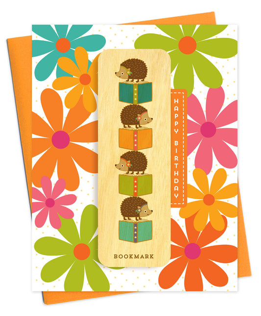 Folded birthday greeting card with bold retro flowers in orange, pink, teal and green, Happy Birthday text and a removable birch wood bookmark featuring hedgehogs stacked on colorful books. Pictured with an orange envelope.