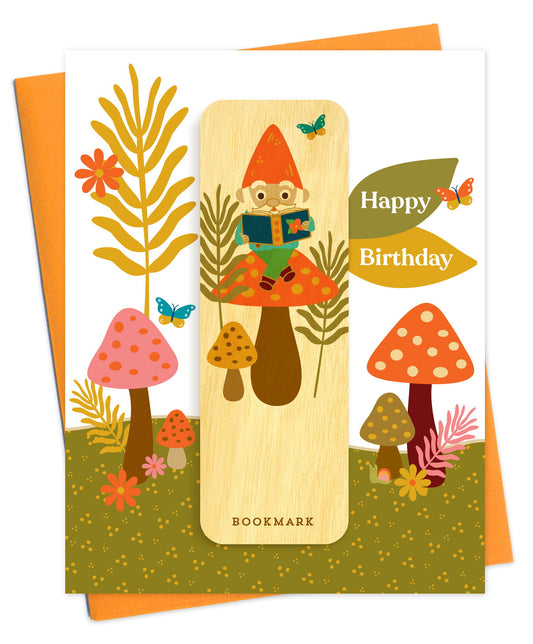Folded birthday greeting card with a whimsical woodland mushroom scene, Happy Birthday text and a removable birch wood bookmark featuring an illustrated gnome reading a book. Pictured with an orange envelope.