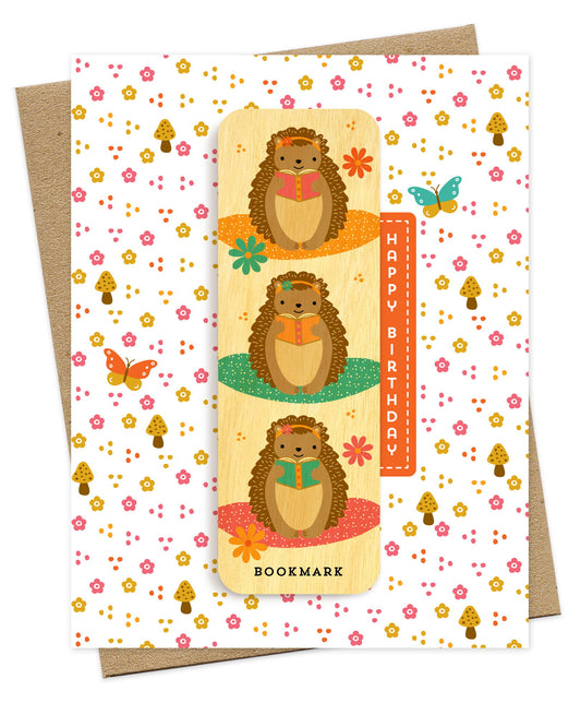 Folded birthday greeting card with a whimsical floral and mushroom pattern, Happy Birthday text and a removable birch wood bookmark featuring illustrated hedgehogs reading books. Pictured with a kraft envelope.