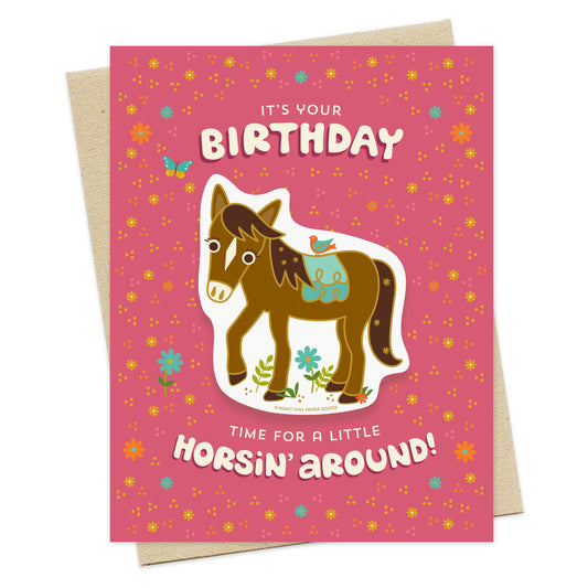 Front of greeting card with a removable horse vinyl sticker and "It's Your Birthday Time for a Little Horsin' Around" text surrounded by small flowers and designs on a pink background. Shown with a light tan envelope.