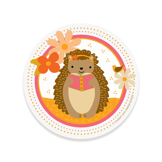 Round vinyl sticker featuring a hedgehog reading a book, surrounded by flowers and a bird on a white background.