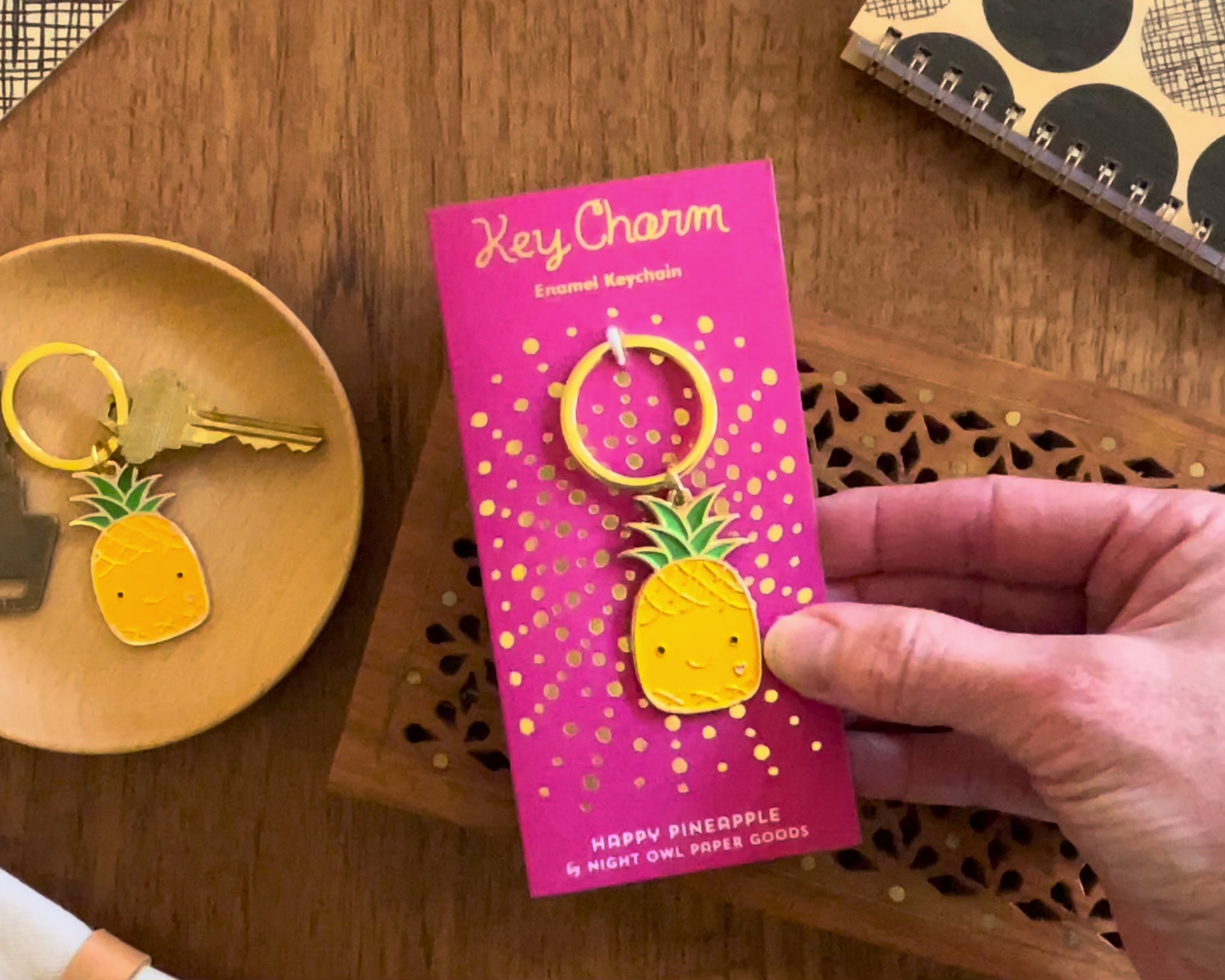 Front of enamel keychain is a smiling pineapple with gold accents. Keychain is mounted on a backer card that says "Key Charm - Enamel Keychain - HAPPY PINEAPPLE by Night Owl Paper Goods" and is packaged in a clear box.