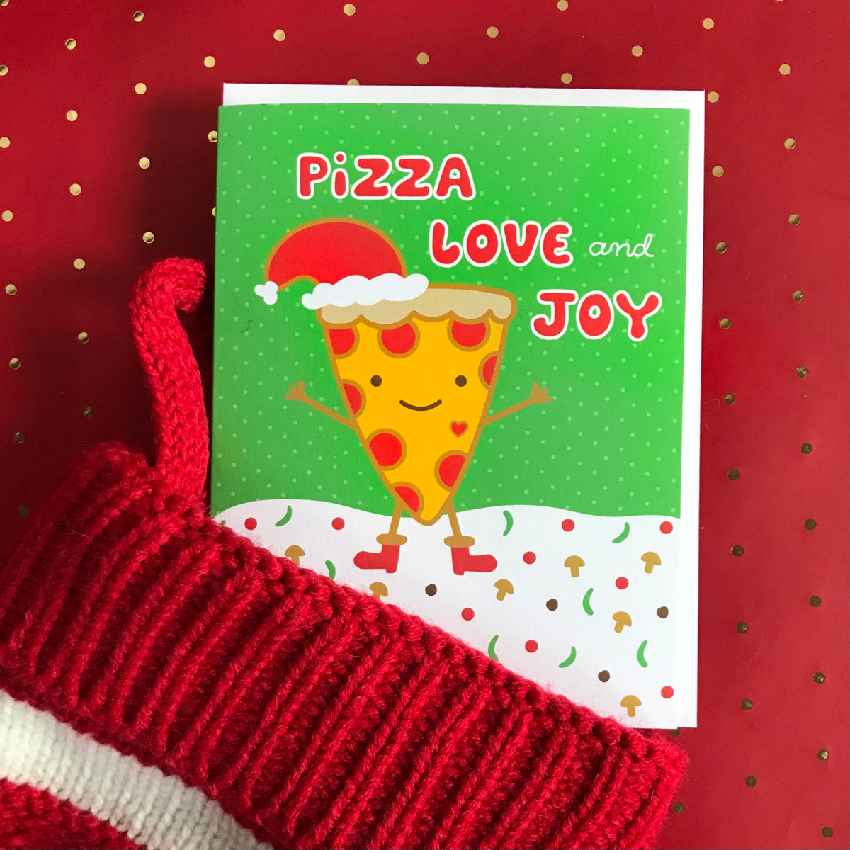 Pizza Love Joy folded paper holiday card featuring a smiling pepperoni pizza slice in a Santa hat and red boots dancing on a snowy ground scattered with mushrooms and peppers against a green background with the greeting "Pizza Love and Joy." Shown emerging from a knit stocking with a white envelope.